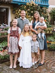 Eine frohe Familie steht in traditioneller Kleidung vor einer blühenden Gartenlandschaft. Sie lächeln und posieren freundlich für das Foto.
