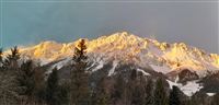 Ein beeindruckendes Bergpanorama mit schneebedeckten Gipfeln im warmen Licht der Abenddämmerung. Die Bäume im Vordergrund ergänzen die natürliche Schönheit der Landschaft.