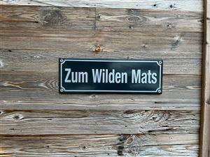 A wooden sign with the inscription "To the Wild Mats". It is attached to a rustic wall.