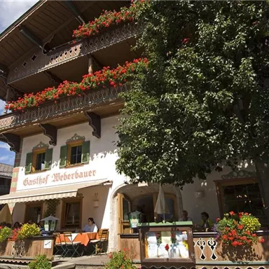 A traditional inn with blooming window boxes and a large tree in the foreground. The sky is clear and the place exudes an inviting atmosphere.