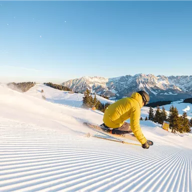 A skier glides over freshly groomed slopes in the snow. In the background, beautiful mountains and a clear sky are visible.