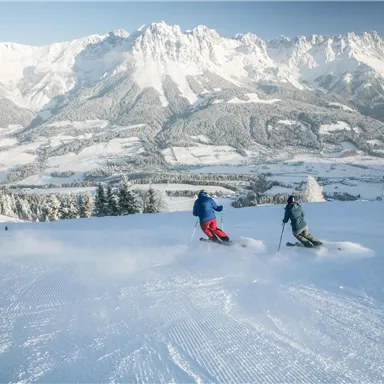 Two skiers glide over a snow-covered slope. In the background, impressive mountains rise under a clear sky.