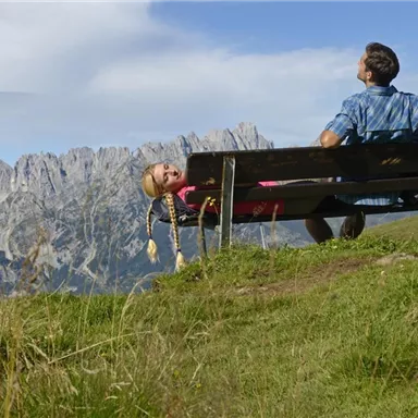 A man is sitting on a bench in the mountains while a girl is lying relaxed next to him. The stunning landscape with tall mountains and green grass can be seen in the background.