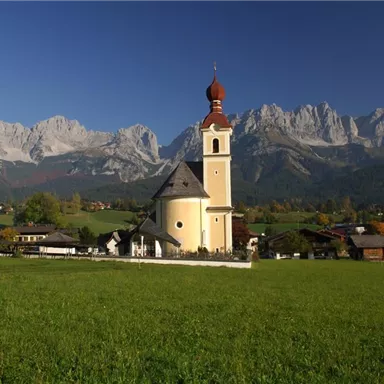 A picturesque church stands in a green meadow, surrounded by impressive mountains. The clear sky and autumn colors give the scene a tranquil atmosphere.
