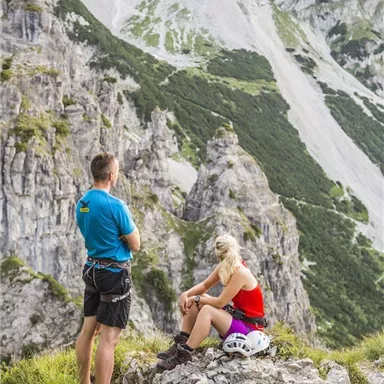 Two mountaineers are standing and sitting on a rock in the mountains. In the background, steep rock walls and a green landscape are visible.
