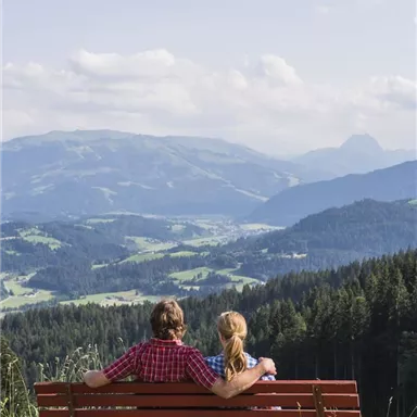 A couple is sitting on a bench, looking at a picturesque mountain landscape. The sky is clear and nature is lush green.