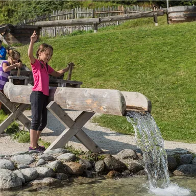 Kinder spielen an einem Wasserspielplatz und experimentieren mit Wasser. Im Hintergrund sind grüne Wiesen und Holzstrukturen zu sehen.