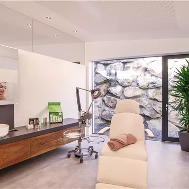 A modern treatment room with bright walls and a large window. The room contains a couch, plants, and an elegant wooden counter.