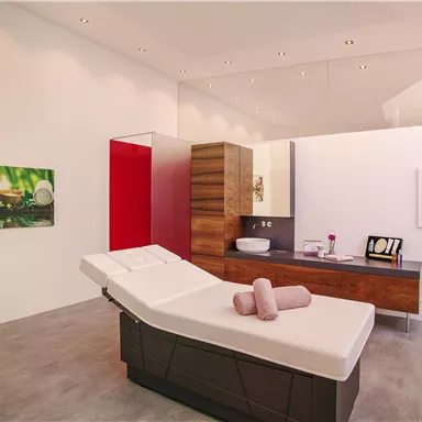 A modern wellness room with a massage bed and wooden shelves. Pictures of relaxing nature motifs hang on the walls.