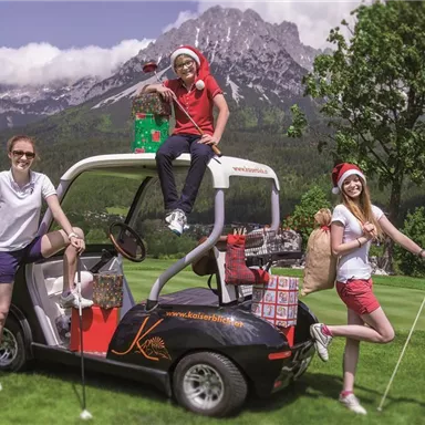 A cheerful golf scene with three people in Christmas hats standing around a golf cart loaded with presents. In the background, mountains and a clear sky are visible.