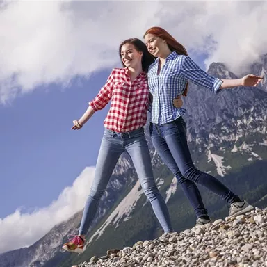 Two cheerful young women are standing on a gravel field and laughing. In the background, majestic mountains and a blue sky with some clouds can be seen.