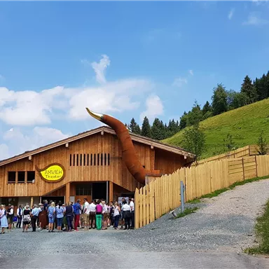 A wooden building with an impressive horn extension, surrounded by green meadows. A large group of people stands in front of the building and enjoys the day.