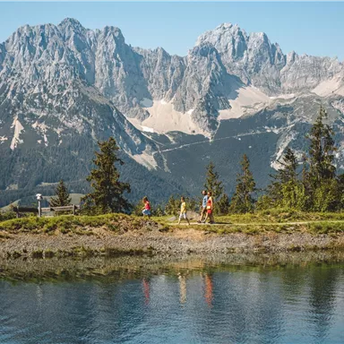 A picturesque mountain landscape with high peaks and clear water in the foreground. A few people are enjoying nature by the shore.