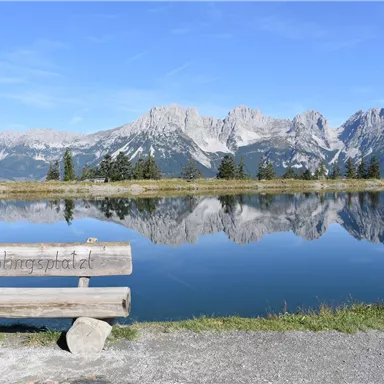 A wooden bench by the calm lake with mountains in the background. The water reflects the surrounding landscape and the clear sky.