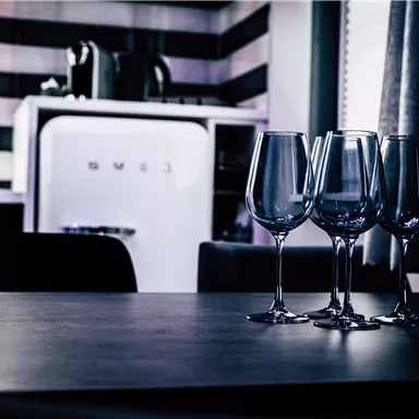 Four wine glasses are on a table in a modern kitchen. In the background, a refrigerator is visible.