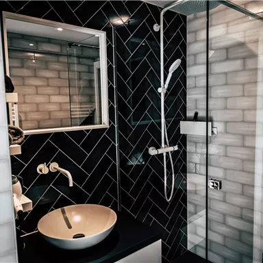 A modern bathroom with a black tiled wall and an elegant shower. There is a round sink and a large mirror.
