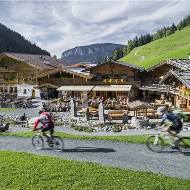 Two cyclists ride along a path past a traditional mountain inn. In the background, green hills and mountains can be seen.