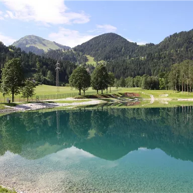 A calm lake surrounded by green meadows and mountains. The water reflects the landscape and the blue sky.