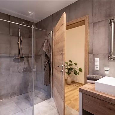 A modern bathroom with a glass shower and a wooden sink. The walls are in concrete gray, and in the background, there is a plant.