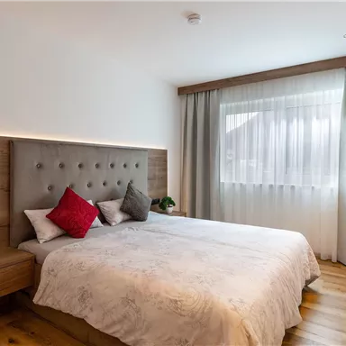 A modern bedroom with a large bed and elegant pillows. Daylight streams through a window with curtains onto the wooden floor.