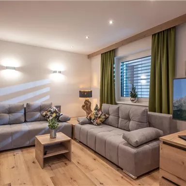 A modern living room with gray sofas and a wooden floor. The windows are decorated with green curtains and a television is placed on a wooden cabinet.