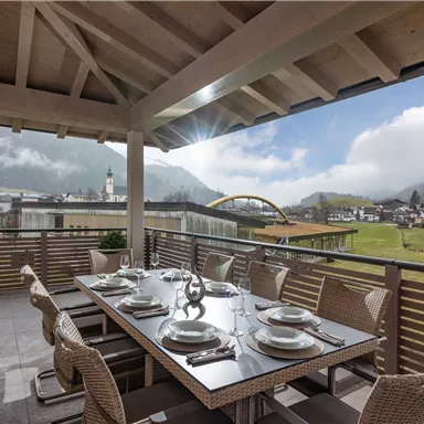 A cozy dining area on a terrace with a view of the mountains. The table is set and the sun is shining through the clouds.
