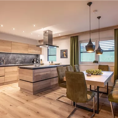 A modern kitchen with wooden surfaces and a cooking island. The dining area is cozy with a wooden table and green chairs.