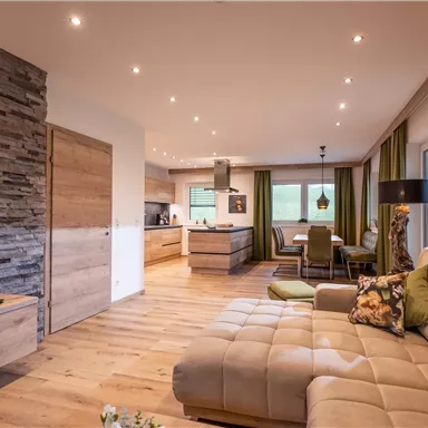 A modern living room with a comfortable sofa and a TV. In the background, there is an open kitchen and large windows overlooking nature.