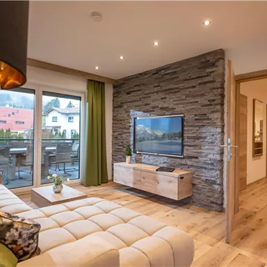 A modern living room with a cozy sofa and a television. Large windows offer a view of a balcony and plenty of daylight.