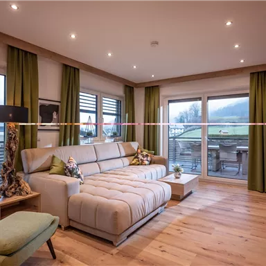 A modern living room with a bright wooden floor, a cozy sofa, and large windows. The green curtains and the view outside create an inviting atmosphere.