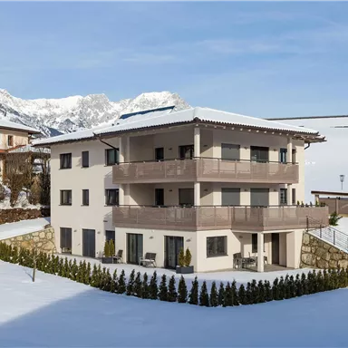 A modern three-story house in the mountains, surrounded by snow. In the background, majestic mountain peaks can be seen.