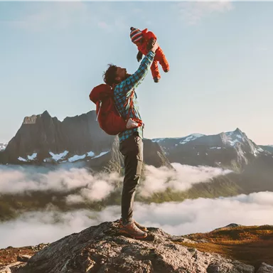 A person lifts a child into the air on a mountain summit. Impressive mountains and clouds can be seen in the background.