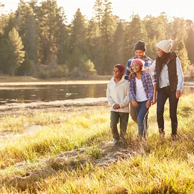 A group of four children stands at the shore of a calm body of water, surrounded by trees. The sun is shining and it is a cheerful atmosphere.