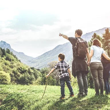 A family is standing in a meadow and looking at the mountains. The man is pointing into the distance while the others are looking attentively.