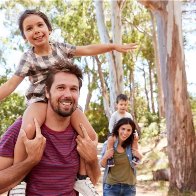 A family is enjoying an outing in nature. A father is carrying his daughter on his shoulders, while the mother and son stand in the background.