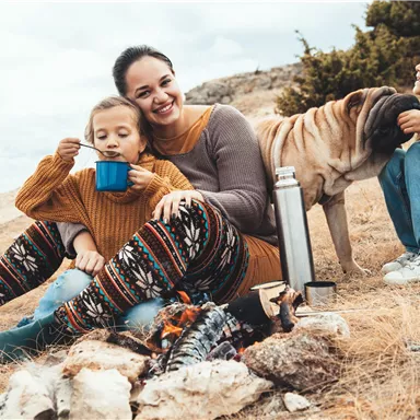 A family is sitting outside by the campfire. They are enjoying hot drinks and have their dog with them.