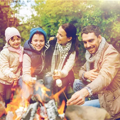 A cheerful group of children and adults sits around a campfire. They laugh and enjoy their time in nature.