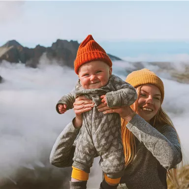 A smiling mother holds a baby in her arms, both wearing colorful hats. In the background, mountains and a blanket of clouds can be seen.