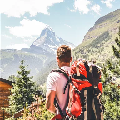 A hiker with a large backpack looks at the impressive mountains. The landscape is green and the clouds are passing by in the sky.