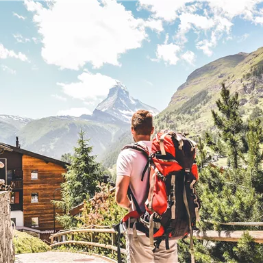 A hiker with a backpack stands in front of an impressive mountain landscape. In the background, the Matterhorn is visible, surrounded by green hills and a clear blue sky.