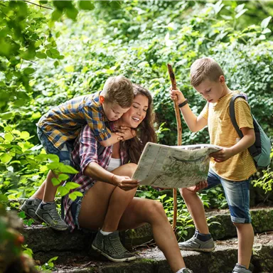 A mother sits in the greenery and looks at a map with her two children. The children appear curious and happy as they explore the path.