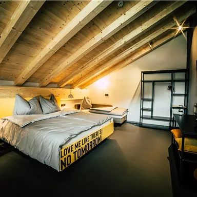 A modern room with sloped ceilings and wooden beams. It features a large bed, a cozy armchair, and stylish decor.