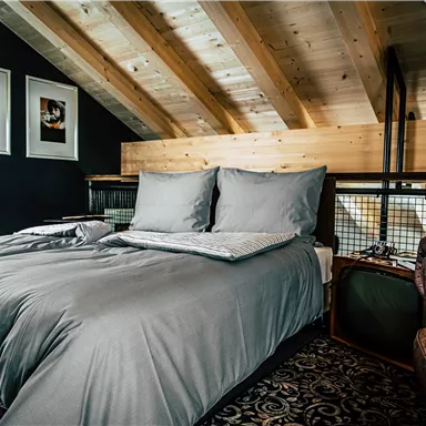 A stylish bedroom with a large bed and a modern brown leather chair. The wooden beam ceiling and the pictures on the wall give the room warmth and coziness.