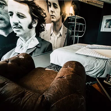 A stylish bedroom with a large wall mural depicting historical musicians. The furnishings include a cozy armchair and a large bed.