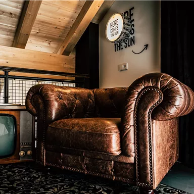 A cozy brown leather armchair is in a modern room. In the background, there is a retro television and a beautiful wooden ceiling.