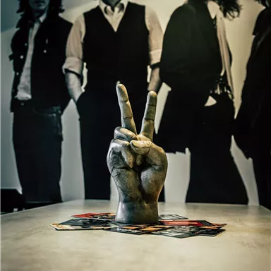 A hand with the peace sign is on a table. In the background, silhouettes of four men can be seen.