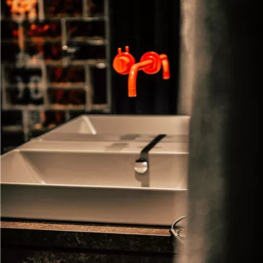 A modern sink with a clean, white design and striking red faucets. Stylish dark tiles are visible in the background.