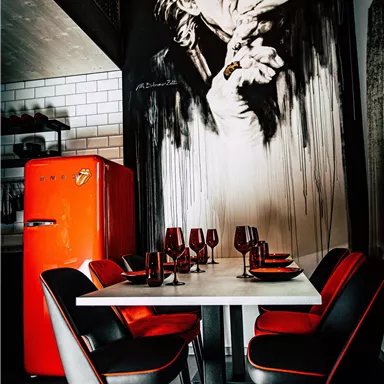 A modern dining room with a white table and black chairs. In the background, there is a large mural and a red refrigerator.