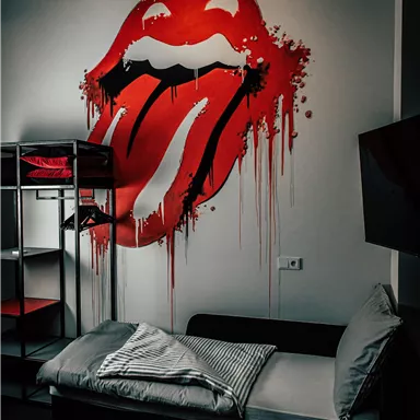 A wall with a striking red lips design. Next to it stands a simple bed with pillows and a blanket.