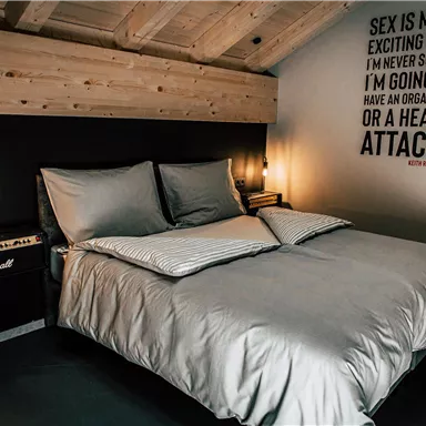 A modern bedroom with a large bed and wooden beams on the ceiling. On the wall is a humorous quote and an amplifier in the corner.